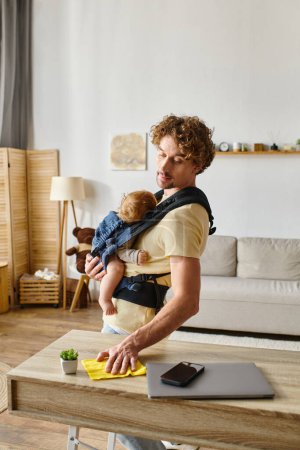 Beschäftigter Vater mit Säugling in Tragetuch-Wischtisch mit gelbem Lappen in der Nähe von Gadgets und winzigen Pflanzen