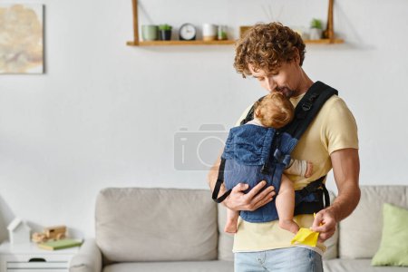 happy father kissing sleeping infant son in carrier and holding yellow rag in modern apartment