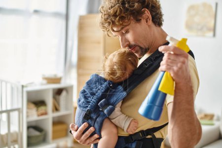 glücklicher Vater küsst verschlafenen Säugling im Tragetuch und hält Sprühflasche und Lappen in der Hand, Hausarbeit