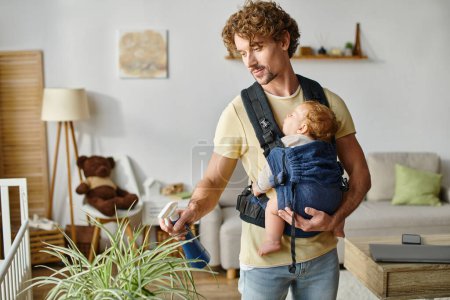 curly father with infant son in carrier holding spray bottle and watering green plant at home