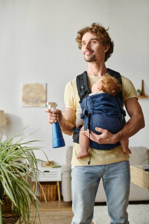 happy father with infant son in carrier holding spray bottle and watering green plant at home