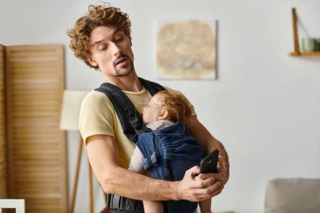 Lockenkopf, alleinerziehender Vater, Nachrichten auf dem Smartphone, während Baby in Tragetasche schläft, moderne Elternschaft