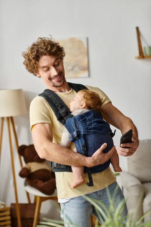 curly haired and happy man messaging on smartphone while baby asleep in carrier, modern parenting