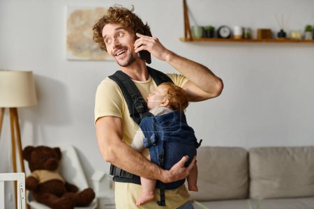 cheerful man talking on smartphone with baby sleeping in carrier, balance between life and work