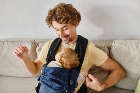 cheerful man with curly hair looking at his infant son in baby carrier, fatherhood and love
