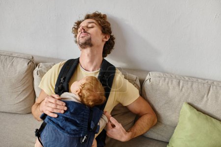 padre de pelo rizado durmiendo con hijo pequeño en portabebés en sala de estar, paternidad y amor