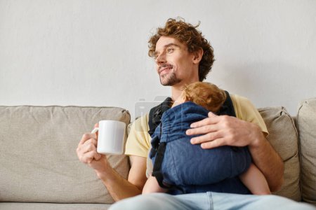 Téléchargez les photos : Père gai avec enfant fils dans le porte-bébé tenant tasse de thé dans le salon, la paternité - en image libre de droit