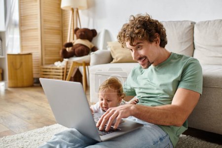 cheerful father using laptop near infant son in living room, balancing between fatherhood and job