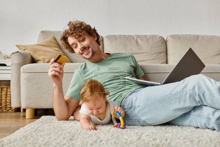 alegre padre sosteniendo tarjeta de crédito mientras que hace compras en línea cerca de niño jugando con sonajero