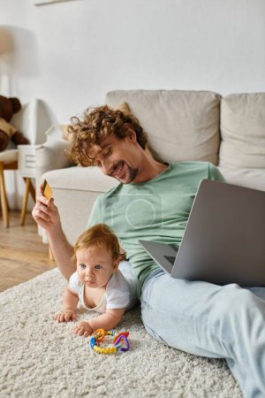 smiling man holding credit card while doing online shopping near baby boy on carpet with rattle