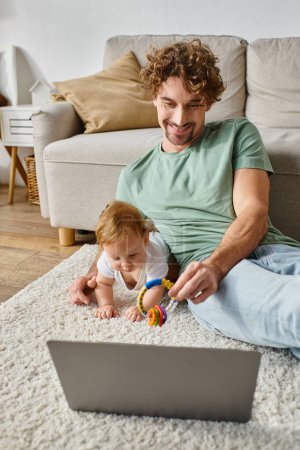 happy man holding baby rattle near infant son and laptop on carpet, balancing between work and life