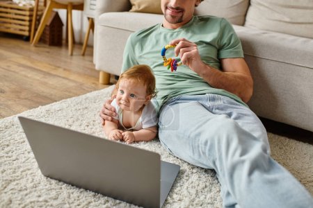 joyful man holding baby rattle near infant son and laptop on carpet, balancing between work and life