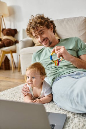 fröhlicher Mann mit Babyrassel in der Nähe des kleinen Sohnes mit blauen Augen und Laptop, Work-Life-Balance