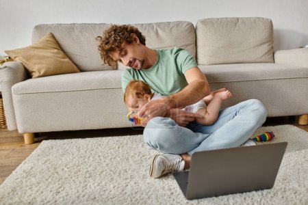 glücklicher Mann mit lockigem Haar hält Babyrassel in der Nähe von Säugling und Laptop, Work-Life-Balance