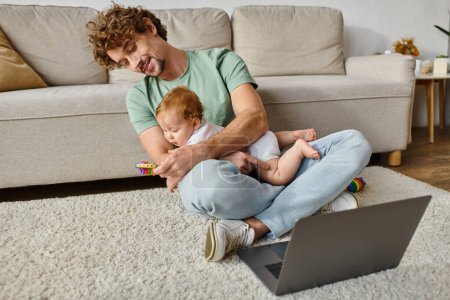 hombre alegre con el pelo rizado que sostiene sonajero del bebé cerca del hijo y del ordenador portátil, trabajo y equilibrio de la vida