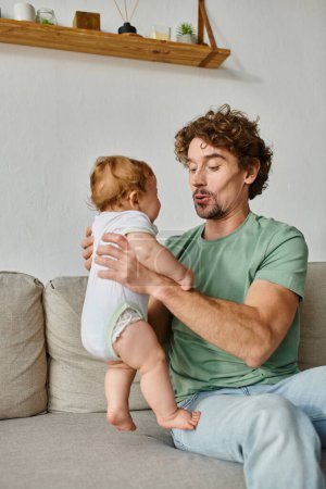 father holding in arms cute infant baby son and having a happy moment on sofa in living room