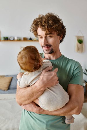 padre cariñoso con pelo rizado y barba sosteniendo en brazos a su niño con ropa de bebé, crianza