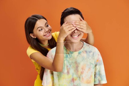 jolly brunette teenage girl closing eyes of her asian friend on orange backdrop, friendship day