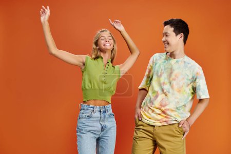 asiatische Teenager-Junge Blick auf seine fröhliche blonde Freundin in lässiger Kleidung auf orangefarbenem Hintergrund
