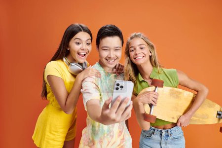 cheerful multiracial teenage friends in vivid attires with headphones and skateboard taking selfie