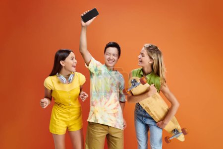 joyous multicultural teenage friends with headphones and skateboard having fun on orange backdrop
