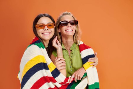 fröhliche blonde und brünette Teenager-Mädchen mit Sonnenbrille, die sich umarmen und in die Kamera schauen, Freundschaft