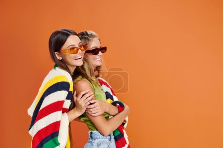 lustige hübsche Teenager-Freunde mit Sonnenbrille, die sich umarmen und mit einer Decke vor orangefarbenem Hintergrund bedecken
