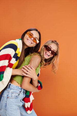 fröhliche entzückende Teenager-Freunde mit Sonnenbrille, die sich umarmen und gemeinsam vor orangefarbener Kulisse Spaß haben