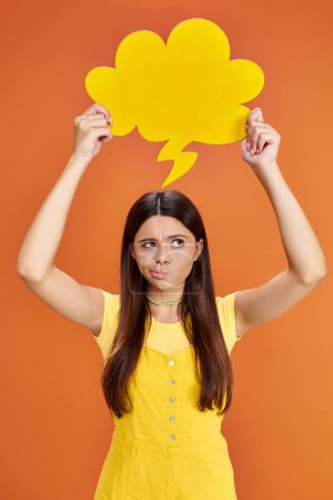 focused adolescent girl in everyday attire holding yellow thought bubble on orange background