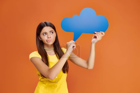 pensive adorable teenage girl in vibrant outfit holding thought bubble on orange background