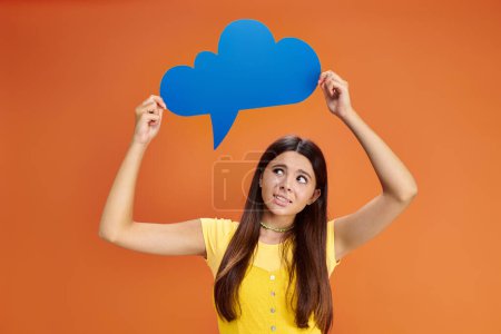 confused teenage girl in casual attire holding thought bubble above her head on orange backdrop