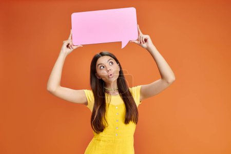 surprised teenage girl in everyday attire holding speech bubble above her head on orange backdrop