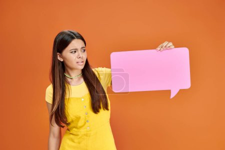 confused pretty adolescent girl in vibrant outfit holding pink speech bubble on orange backdrop