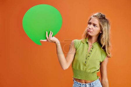 confusa chica adolescente rubia en traje casual mirando la burbuja del habla verde sobre fondo naranja