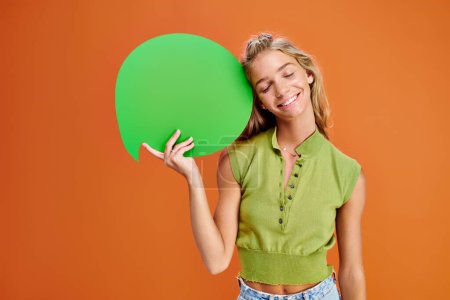 Photo for Joyous teen in casual attire holding speech bubble and posing with closed eyes on orange backdrop - Royalty Free Image