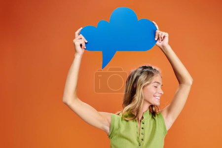jolly blonde teenage girl in casual outfit holding blue thought bubble above head on orange backdrop