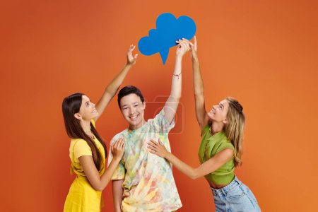 joyous teenage girls reaching blue thought bubble in hands of asian cheerful boy on orange backdrop
