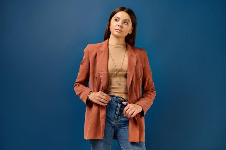 adorable fashionable teenage girl in brown blazer posing on blue backdrop and looking away