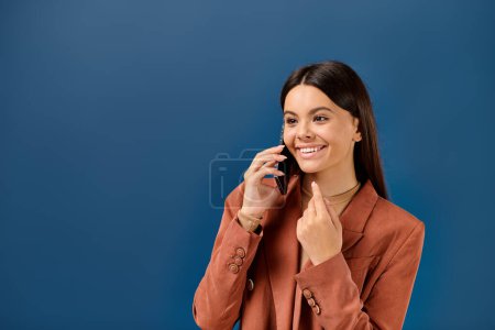 fröhliche stilvolle Teenager-Mädchen im trendigen Blazer telefoniert und lächelt glücklich auf blauem Hintergrund