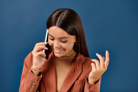 fröhliche brünette Teenager-Mädchen in modischer Kleidung telefoniert und lächelt auf blauem Hintergrund