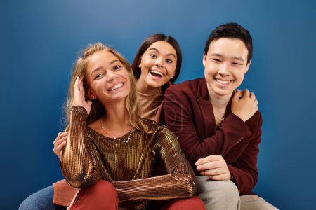 joyful multiracial adolescents in stylish outfits smiling at camera on blue backdrop, friendship day