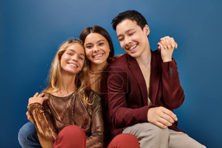 joyful teenage girls smiling at camera next to their jolly elegant asian friend on blue background