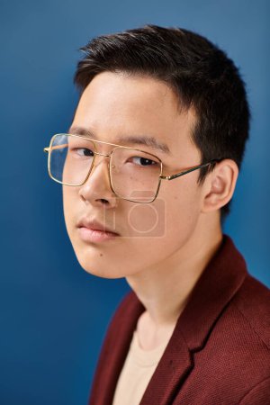 Téléchargez les photos : Bonne recherche asiatique adolescent garçon avec lunettes dans élégant tenue en regardant caméra sur fond bleu - en image libre de droit