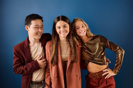jolly multiracial adolescent friends in vivid attires posing on blue backdrop and smiling joyfully