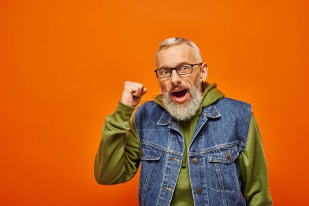 joyful mature funky man in green vibrant hoodie posing with fists near face on orange background