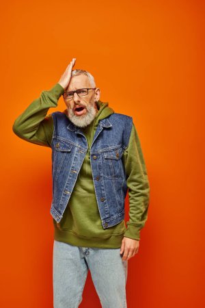 frustrated mature male model in green hoodie and vest looking at camera with hand near face