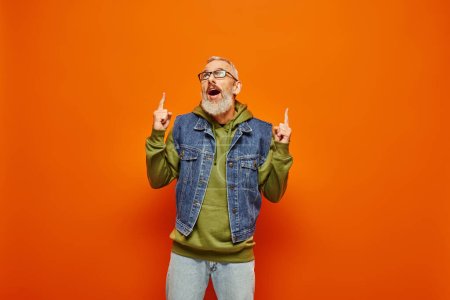 shocked mature man with glasses and gray beard in vivid hoodie pointing up on orange backdrop