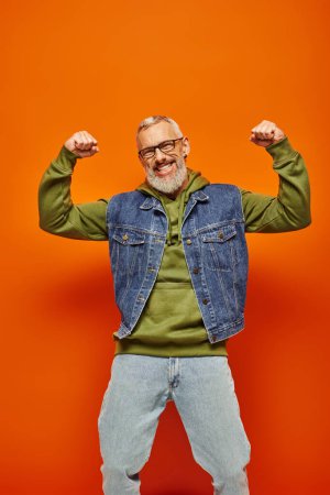 cheerful good looking mature man in denim vest posing with arms raised and smiling at camera