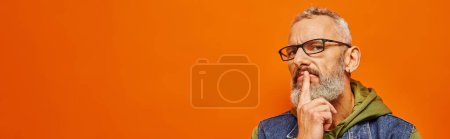 Photo for Focused mature man in green hoodie with glasses and gray beard posing on orange background, banner - Royalty Free Image