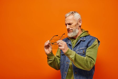 Foto de Guapo hombre maduro concentrado en traje vibrante sosteniendo sus gafas en las manos sobre fondo naranja - Imagen libre de derechos